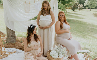 Image shows 3 pregnant women at a picnic. Two are sitting and one is standing. They are all wearing neutral beige dresses.