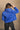 back view of model wearing the Ember Blue Corduroy Puffer Jacket that has royal blue corduroy fabric with a quilted design, pockets, a hidden zipper, a high neck, a drawstring, and long sleeves.