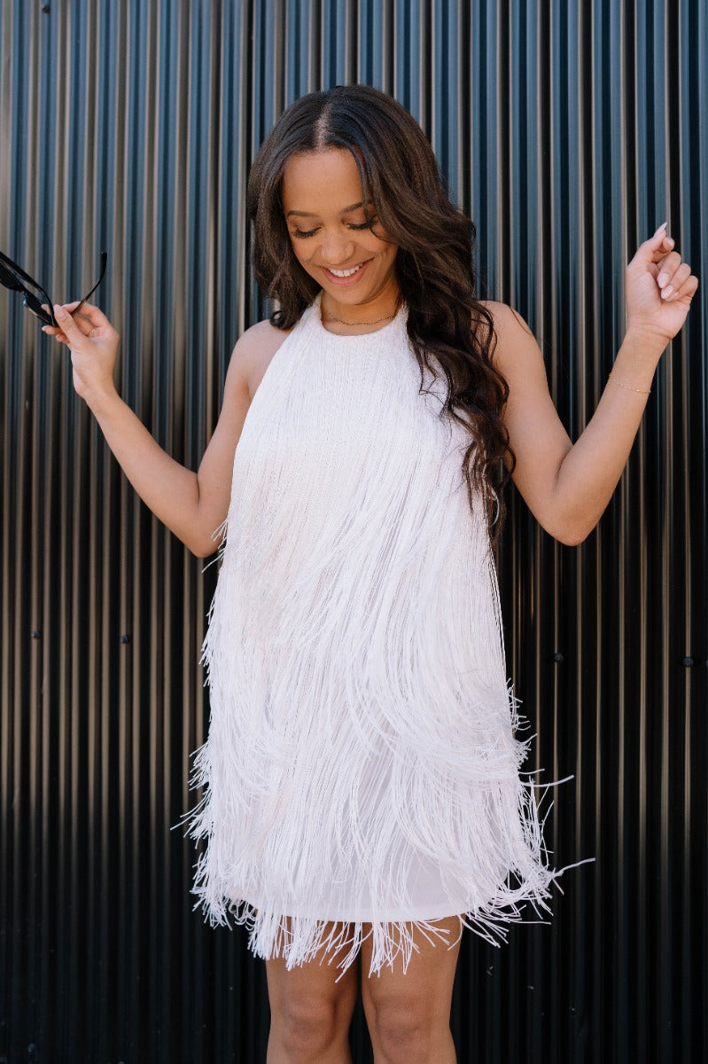 Front view of model wearing the Karissa White Fringe Tiered Mini Dress which features white fringe fabric, white lining, mini length, halter neckline with button closure, sleeveless and open back.