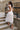 Full body back view of model wearing the Eliana White Floral Midi Dress which features white sheer net fabric, white floral 3D pattern, midi length, white lining, overlap plunge neckline, thick straps with tie details, sleeveless and monochrome back zippe