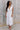 Full body view of model wearing the Eliana White Floral Midi Dress which features white sheer net fabric, white floral 3D pattern, midi length, white lining, overlap plunge neckline, thick straps with tie details, sleeveless and monochrome back zipper wit