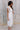 Side view of model wearing the Eliana White Floral Midi Dress which features white sheer net fabric, white floral 3D pattern, midi length, white lining, overlap plunge neckline, thick straps with tie details, sleeveless and monochrome back zipper with hoo