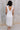 Full body back view of model wearing the Eliana White Floral Midi Dress which features white sheer net fabric, white floral 3D pattern, midi length, white lining, overlap plunge neckline, thick straps with tie details, sleeveless and monochrome back zippe