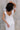 Close up back view of model wearing the Eliana White Floral Midi Dress which features white sheer net fabric, white floral 3D pattern, midi length, white lining, overlap plunge neckline, thick straps with tie details, sleeveless and monochrome back zipper