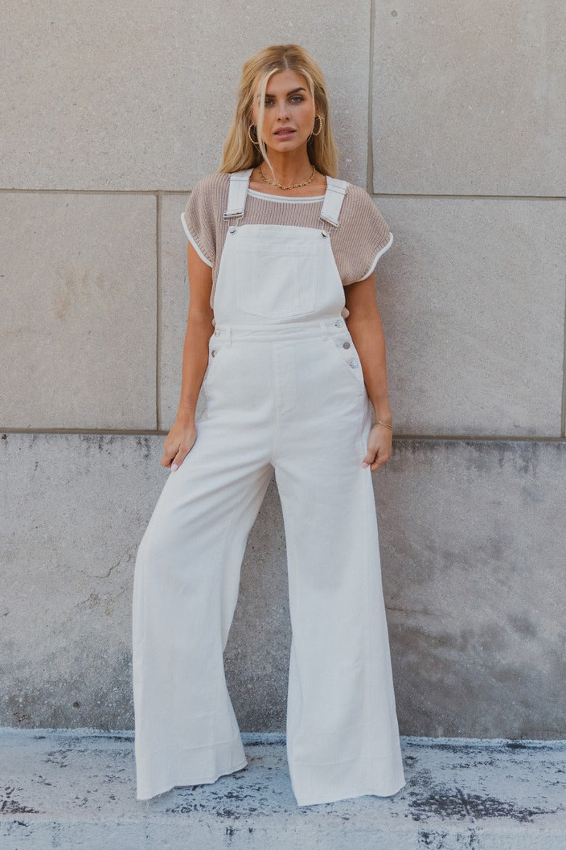 Full body view of model wearing the Bella White Denim Flare Overalls which features white denim fabric, two side slit pockets, belt loops, three button closures on each side, one large front chest pocket, adjustable straps, sleeveless and wide pant legs w