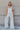 Full body view of model wearing the Bella White Denim Flare Overalls which features white denim fabric, two side slit pockets, belt loops, three button closures on each side, one large front chest pocket, adjustable straps, sleeveless and wide pant legs w
