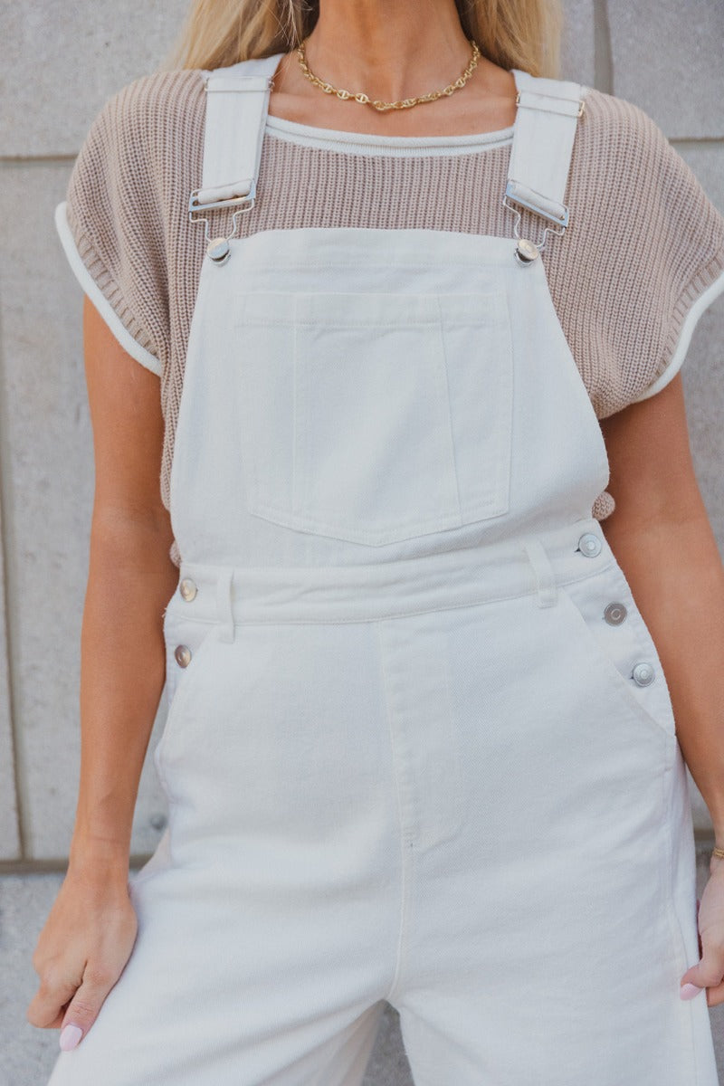 Close up view of model wearing the Bella White Denim Flare Overalls which features white denim fabric, two side slit pockets, belt loops, three button closures on each side, one large front chest pocket, adjustable straps, sleeveless and wide pant legs wi