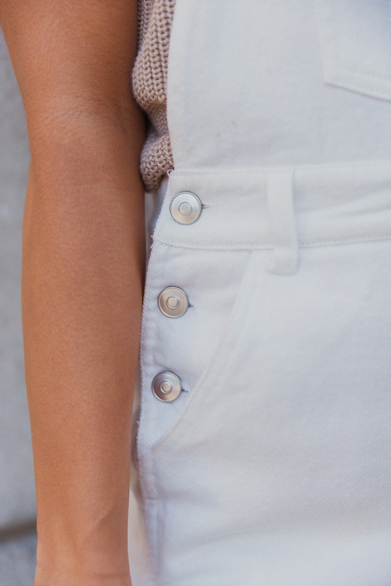 Close up view of model wearing the Bella White Denim Flare Overalls which features white denim fabric, two side slit pockets, belt loops, three button closures on each side, one large front chest pocket, adjustable straps, sleeveless and wide pant legs wi