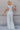 Back view of model wearing the Bella White Denim Flare Overalls which features white denim fabric, two side slit pockets, belt loops, three button closures on each side, one large front chest pocket, adjustable straps, sleeveless and wide pant legs with f