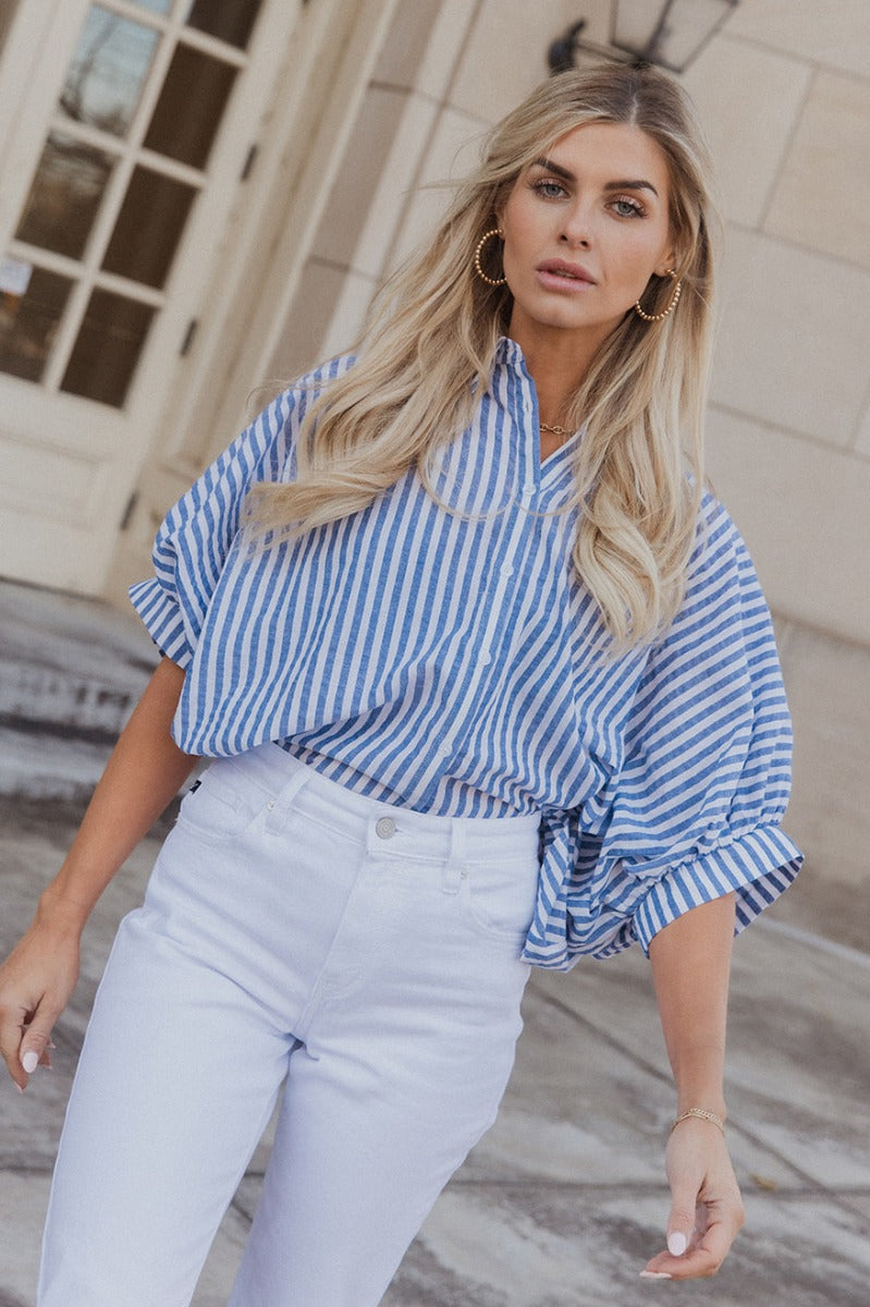 Front view of model wearing the Ella Blue & White Stripe Button Up Top which features dark blue and white fabric with a striped pattern, white button-up front closures, an oversized fit, a collared neckline and short sleeves with cuffs.