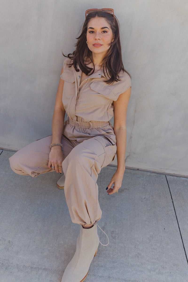 Full body view of model wearing the Zoe Khaki Sleeveless Cargo Jumpsuit which features washed denim fabric, mini length, a small slit detail, a monochrome side zipper with a hook closure, and a monochrome adjustable belt with a buckle.