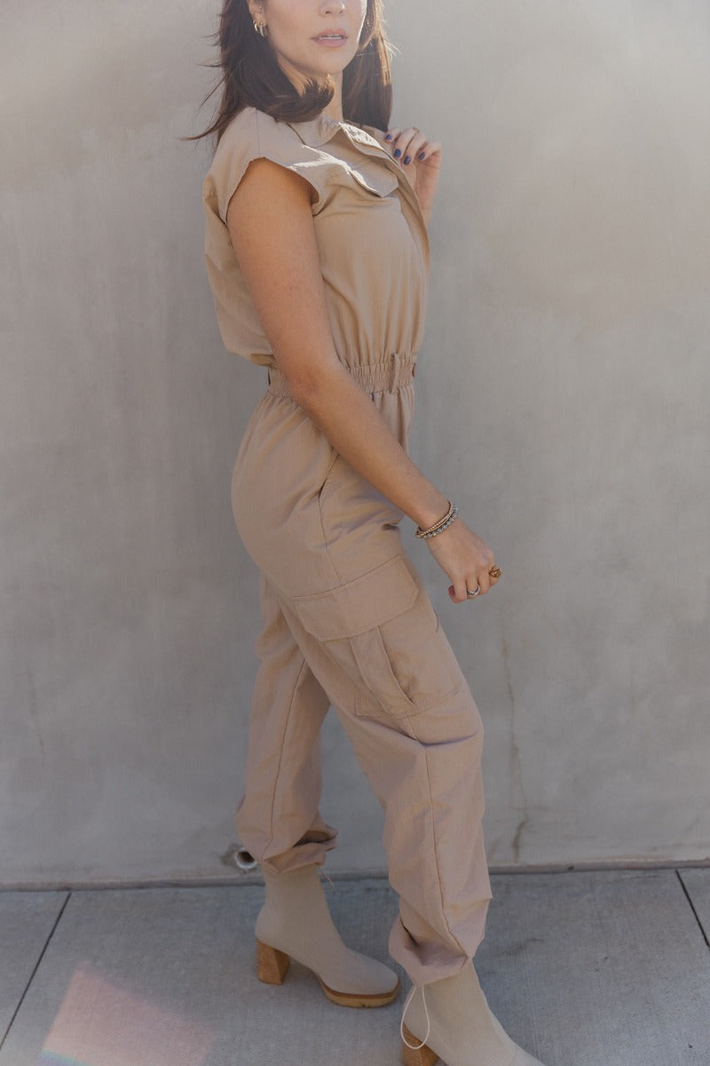 Full body side view of model wearing the Zoe Khaki Sleeveless Cargo Jumpsuit which features washed denim fabric, mini length, a small slit detail, a monochrome side zipper with a hook closure, and a monochrome adjustable belt with a buckle.