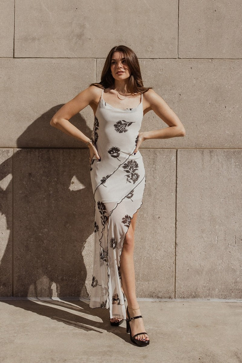 Full body front view of model wearing the Delaney Cream & Black Floral Asymmetric Mesh Midi Dress that has cream mesh fabric with black floral print, black ruffle trim, asymetrical hem with slits, a cowl neck, and thin straps.