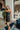 Full body front view of model wearing the Big Reputation Dress, which features a black faux-leather material, a surplice, sweetheart neckline, a sleeveless design with thin adjustable straps, a draped front, a surplice bottom, and a zip-up back closure