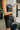 Close-up side view of model wearing the Big Reputation Dress, which features a black faux-leather material, a surplice, sweetheart neckline, a sleeveless design with thin adjustable straps, a draped front, a surplice bottom, and a zip-up back closure