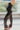 Side view of model wearing The Hailey Jumpsuit features black faux-leather, a sleeveless design, a straight neckline, a tie belt at the waist, a back zipper closure, and a straight leg fit.