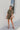 Frontal view of the The Lady May Romper that features an olive green fabric, a collar neckline, a short cuffed sleeve, a button-up front, two front pockets at the chest, a cuffed bottom hem, and two back pockets.