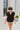 Frontal view of the Irreplaceable Top that features a black colored material, a round neckline with a ruffle detail, a tie detail with a cut-out at the chest, a short sleeve, and a fowy fit