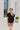 Frontal view of the Irreplaceable Top that features a black colored material, a round neckline with a ruffle detail, a tie detail with a cut-out at the chest, a short sleeve, and a fowy fit
