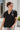 Frontal view of the Irreplaceable Top that features a black colored material, a round neckline with a ruffle detail, a tie detail with a cut-out at the chest, a short sleeve, and a fowy fit