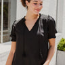 Frontal view of the Irreplaceable Top that features a black colored material, a round neckline with a ruffle detail, a tie detail with a cut-out at the chest, a short sleeve, and a fowy fit