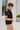 Side view of the Irreplaceable Top that features a black colored material, a round neckline with a ruffle detail, a tie detail with a cut-out at the chest, a short sleeve, and a fowy fit