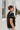Side view of the Irreplaceable Top that features a black colored material, a round neckline with a ruffle detail, a tie detail with a cut-out at the chest, a short sleeve, and a fowy fit