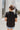 Back view of the Irreplaceable Top that features a black colored material, a round neckline with a ruffle detail, a tie detail with a cut-out at the chest, a short sleeve, and a fowy fit