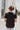 Back view of the Irreplaceable Top that features a black colored material, a round neckline with a ruffle detail, a tie detail with a cut-out at the chest, a short sleeve, and a fowy fit