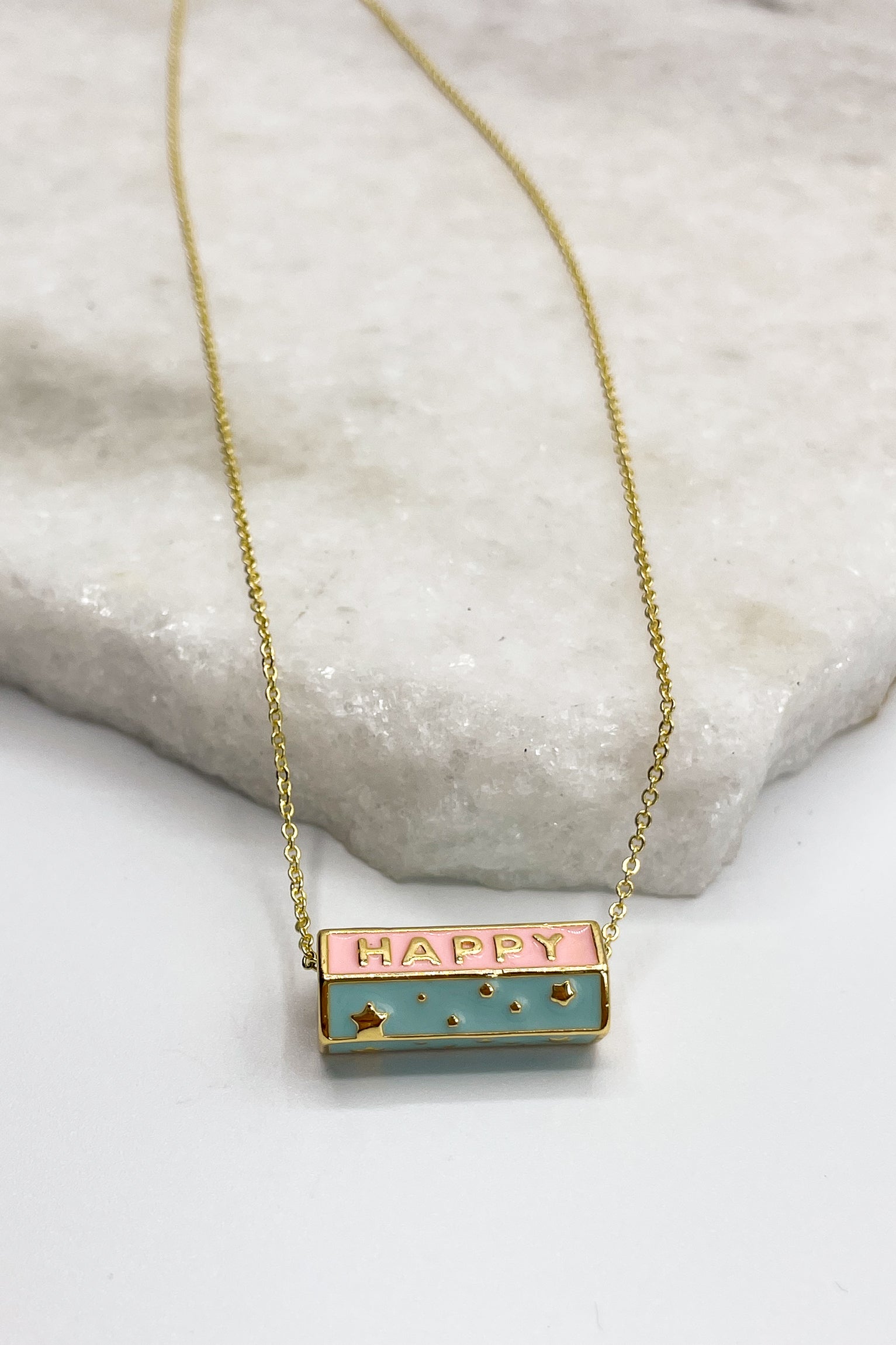 Close up of Happy Gold Necklace, that is a adjustable gold chain link with a blue and pink medallion says HAPPY.