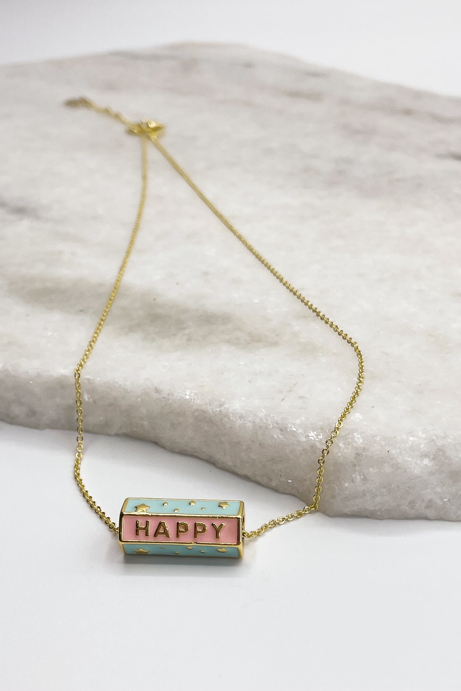 Close up of the medallion on Happy Gold Necklace, that is a adjustable gold chain link with a blue and pink medallion says HAPPY.