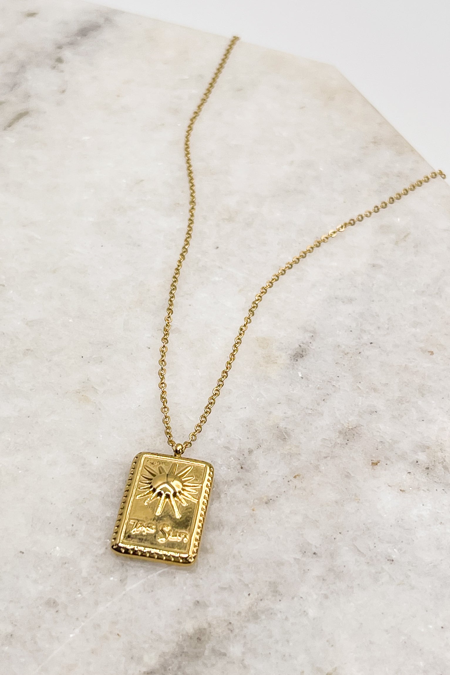 flat lay view of the Orion Sun Gold Necklace which features adjustable gold chain link with rectangle medallion. the medallion says "the sun" with a sun detail