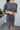 Close-up frontal side view of model wearing the Keep Your Cool Dress, which has a grey ribbed material, a round neck, long sleeves, a front knot detail, a front slit, and a high-low silhouette.