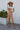 Full body front view of model wearing the Next Adventure Jumpsuit in Camel features a linen material, a square neck, a sleeveless design with tie straps, two front and back pockets, and elastic ankles. Worn over white tshirt.