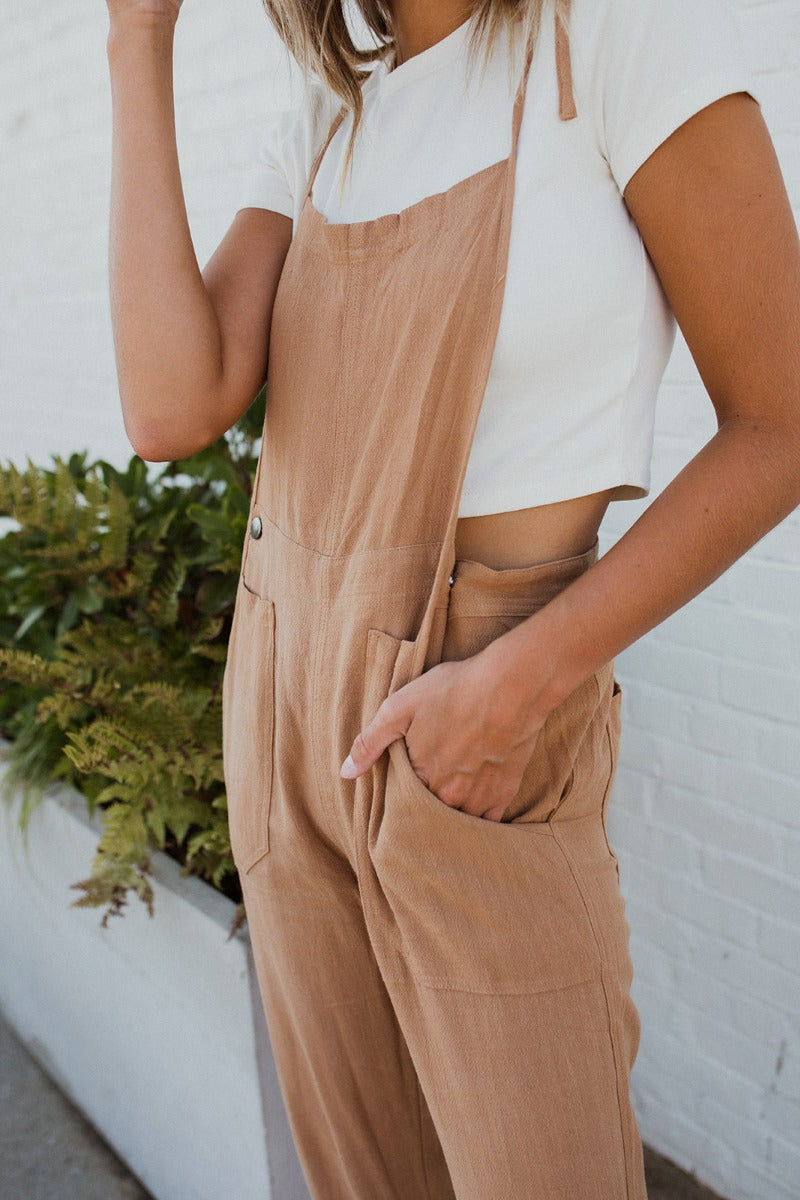 Close-up side view of model wearing the Next Adventure Jumpsuit in Camel features a linen material, a square neck, a sleeveless design with tie straps, two front and back pockets, and elastic ankles. Worn over white tshirt.