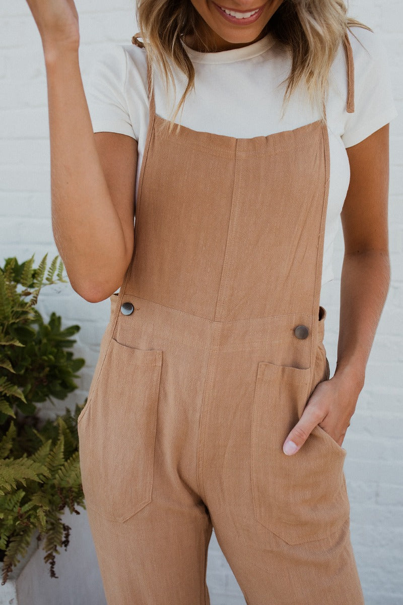 Close-up front view of model wearing the Next Adventure Jumpsuit in Camel features a linen material, a square neck, a sleeveless design with tie straps, two front and back pockets, and elastic ankles. Worn over white tshirt.