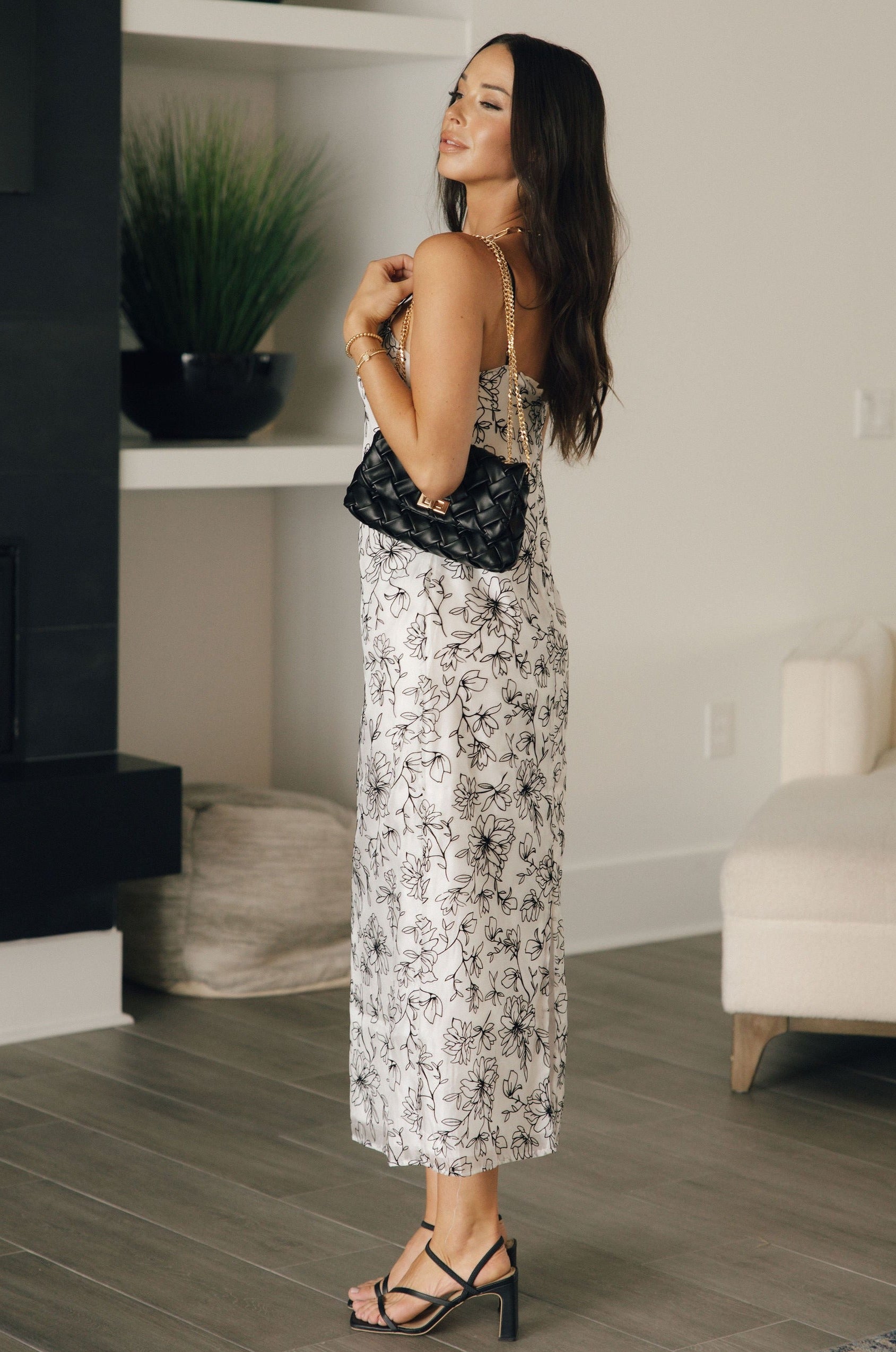 Full body side view of female model wearing the Lauren White & Black Floral Midi Dress which features White Sheer Fabric, Black Floral Print, White Lining, Midi Length, Slit Side Detail, Square Neckline, Black Adjustable Straps and Back Zipper with Hook Closure
