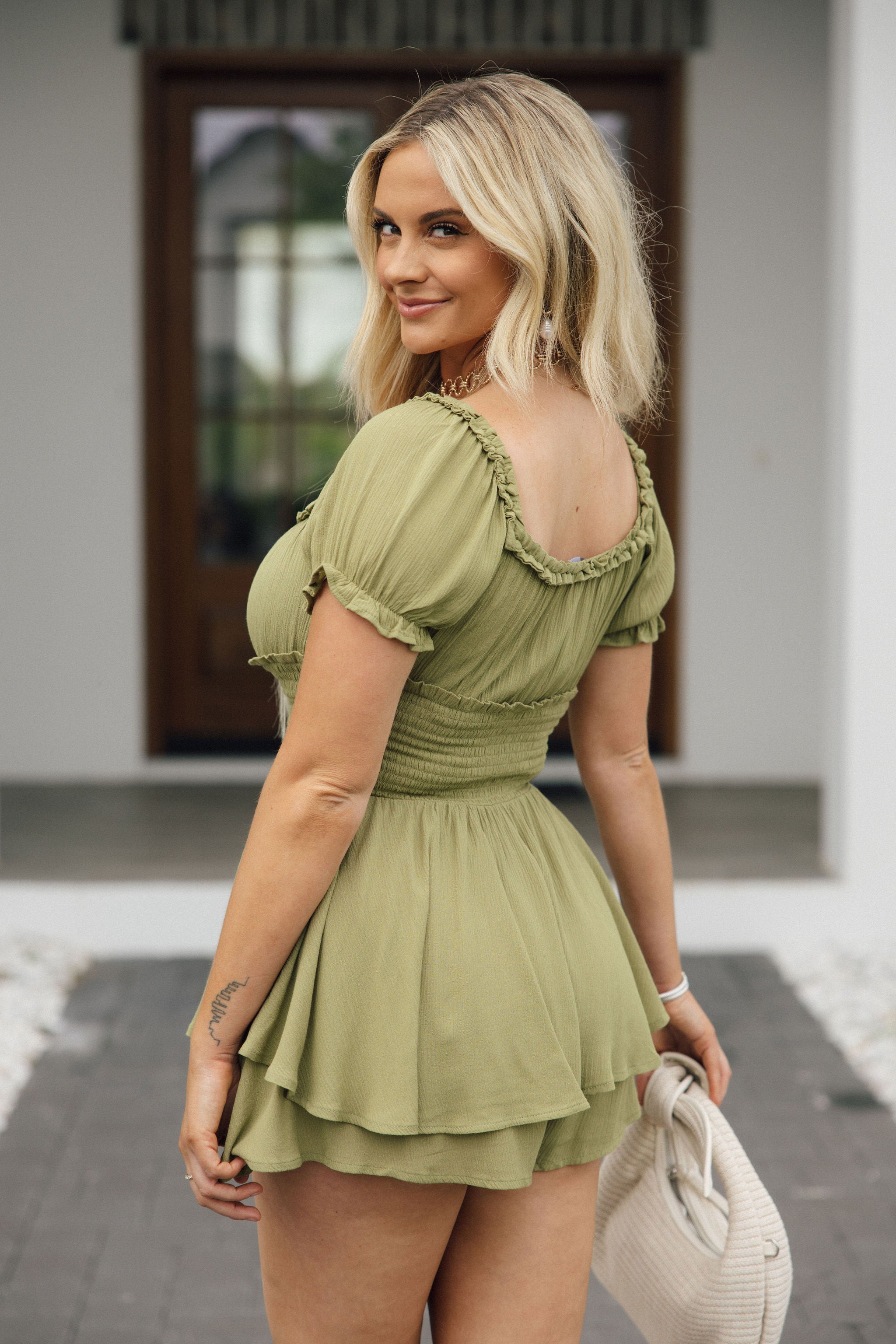 Back view of female model wearing the Natalia Moss Green Ruffle Short Sleeve Romper which features Olive Green Lightweight Fabric, Ruffle Tiered Shorts, Smocked Waistband, Ruffle Hem Details, Square Neckline and short Sleeves