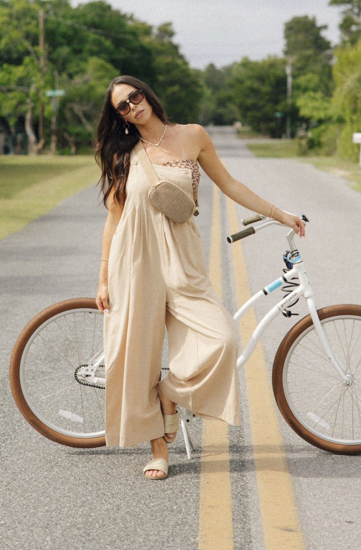 Full body front view of female model wearing the Calista Tan Wide Leg Jumpsuit that has tan fabric, straps that button, and wide legs. Worn over tube top. Model is standing in front of bike.
