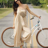 Full body front view of female model wearing the Calista Tan Wide Leg Jumpsuit that has tan fabric, straps that button, and wide legs. Worn over tube top. Model is standing in front of bike.