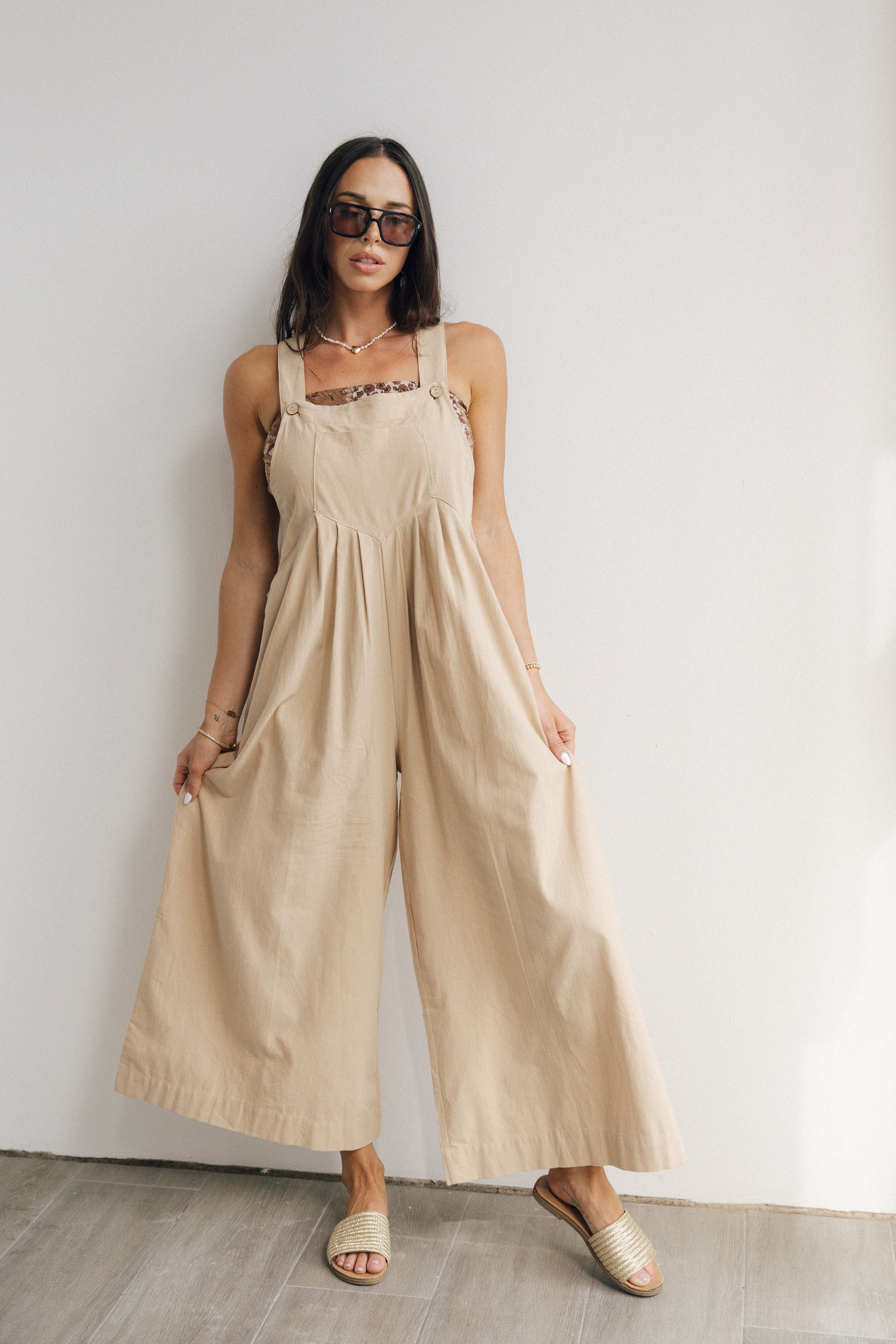 Full body front view of female model wearing the Calista Tan Wide Leg Jumpsuit that has tan fabric, straps that button, and wide legs. Worn over tube top.