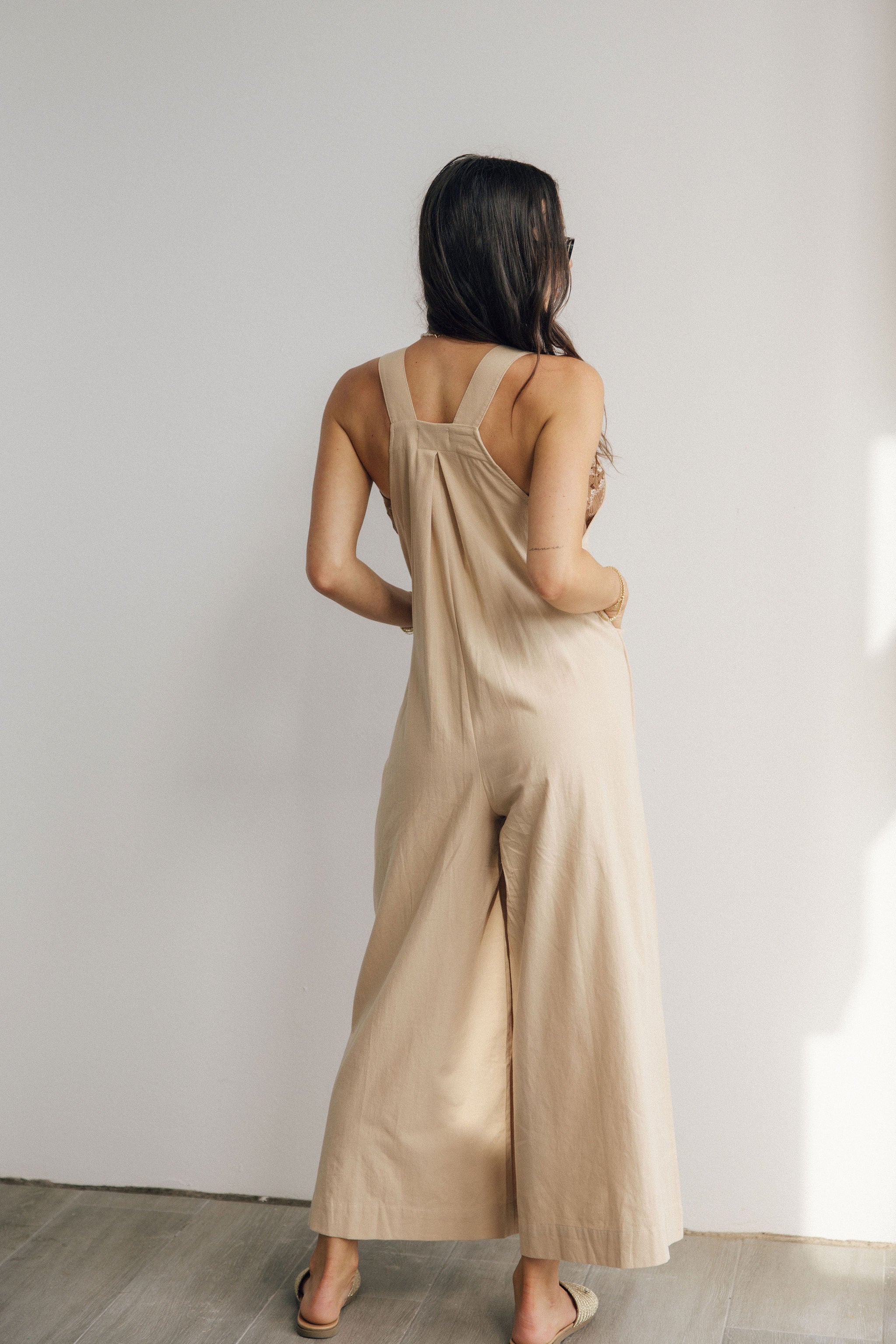 Full body back view of female model wearing the Calista Tan Wide Leg Jumpsuit that has tan fabric, straps that button, and wide legs. Worn over tube top.
