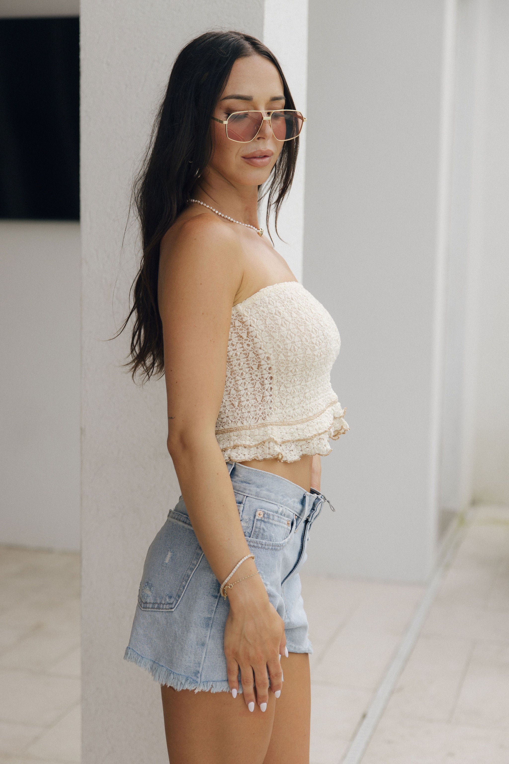 Upper body side view of model wearing the Adley Light Wash Frayed Denim Shorts that have light wash denim, pockets, a frayed hem, and light distressing. Worn with tube top.