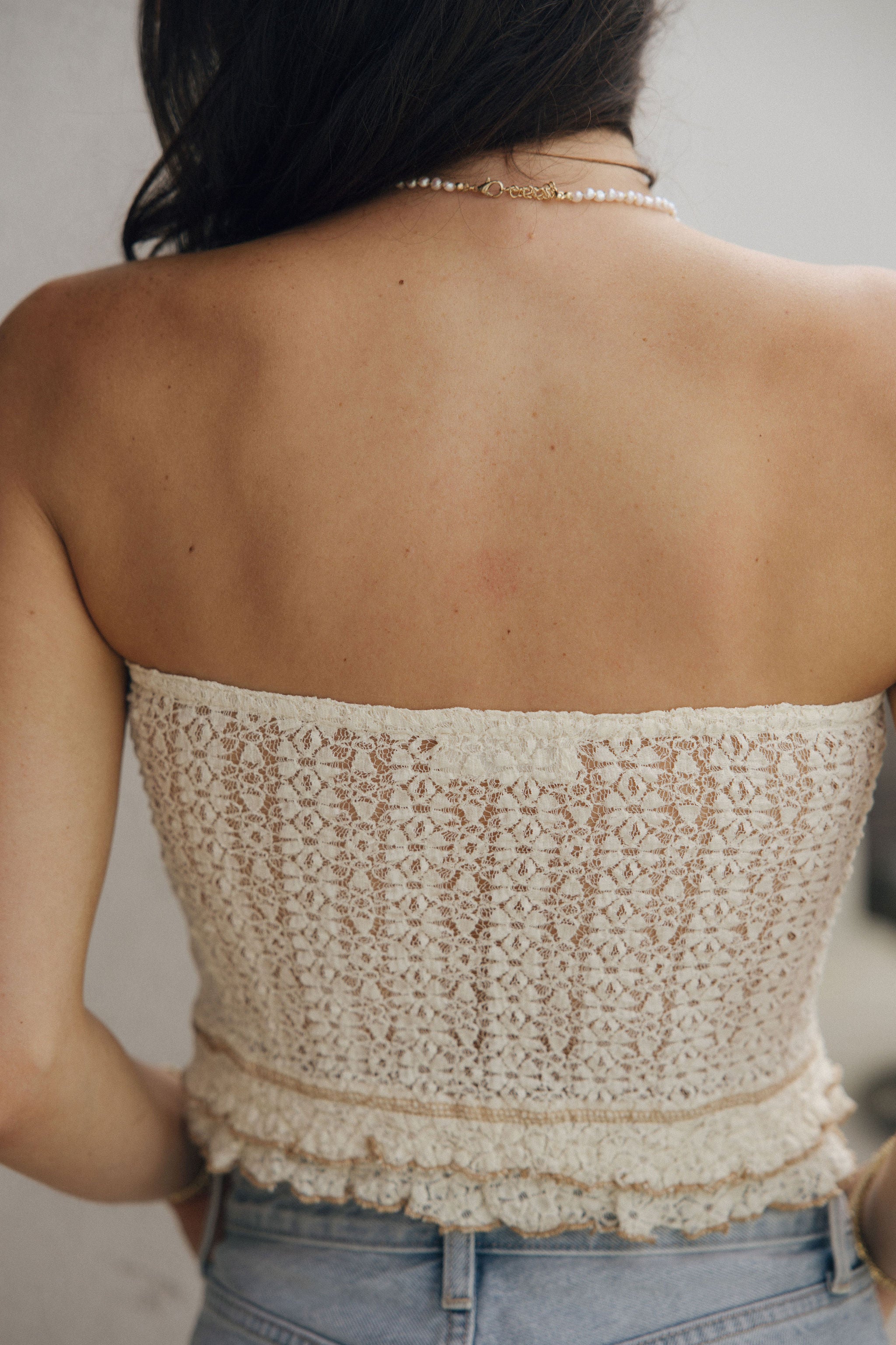 Close up upper body back view of female model wearing the Kyra Cream Lace Tube Top that has cream lace, taupe hem details, and a strapless neck.