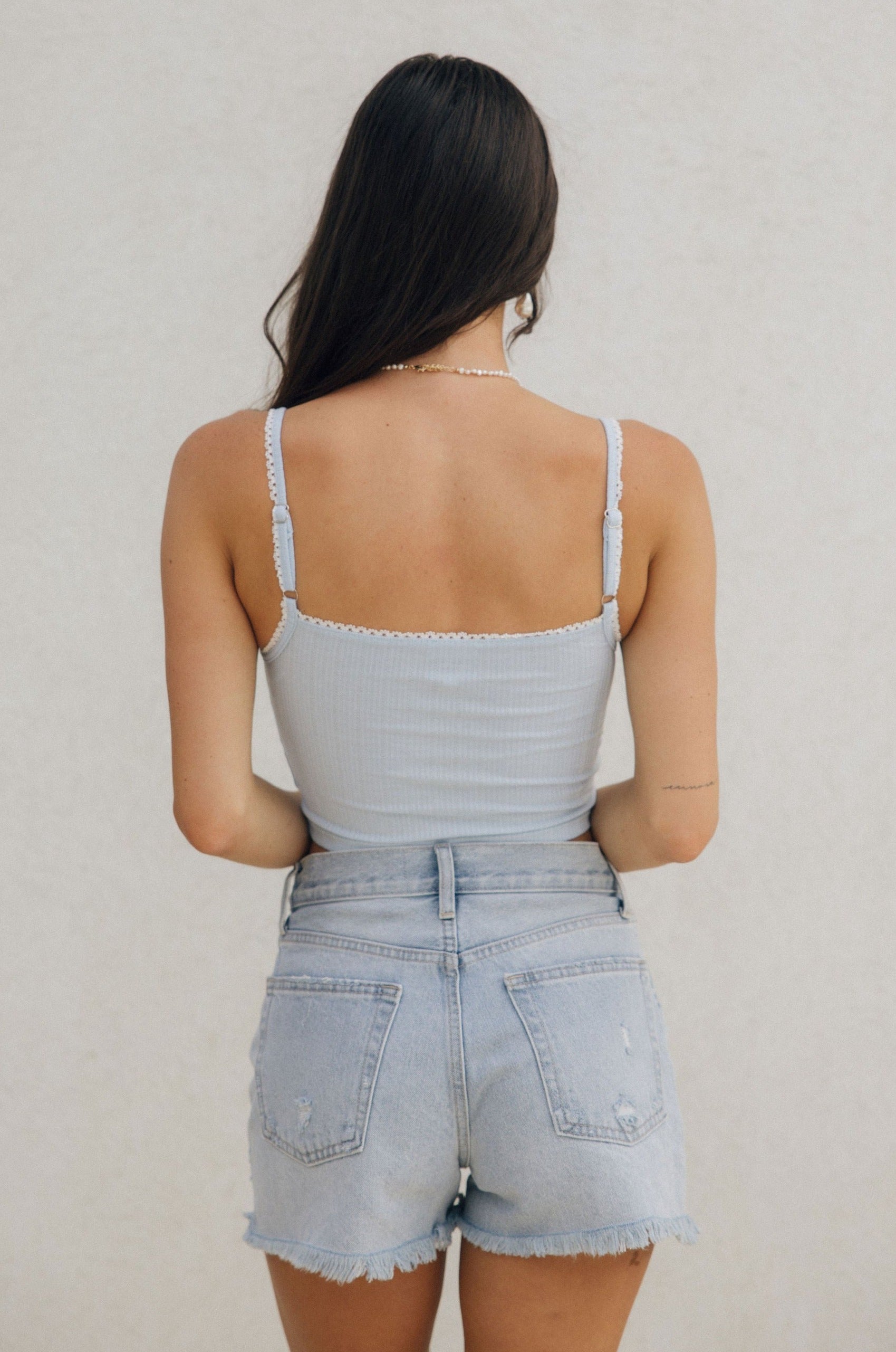Back view of female model wearing the Amora Light Blue Lace Cropped Tank which features Light Blue Ribbed Fabric, White Lace Trim Details, Square Neckline, Adjustable Straps and Cropped Waist