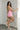 Full body side view of female model wearing the Palmer Pink Ruffle Halter Sleeveless Romper which features Pink Lightweight Fabric, Pink Shorts Lining, Surplice Neckline with Halter Neck Ties, Ruffle Hem Details, Open Back and Back Zipper with Hook Closure