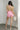 Full body back view of female model wearing the Palmer Pink Ruffle Halter Sleeveless Romper which features Pink Lightweight Fabric, Pink Shorts Lining, Surplice Neckline with Halter Neck Ties, Ruffle Hem Details, Open Back and Back Zipper with Hook Closure