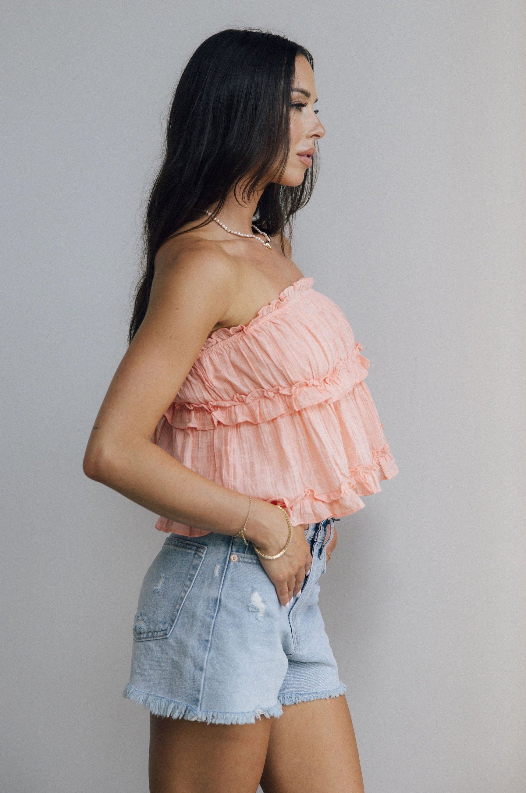 Side view of female model wearing the Millie Peach Ruffle Strapless Top which features Peach Cotton Fabric, Ruffle Tiered Body Details, Peach Lining, Cropped Waist and Strapless