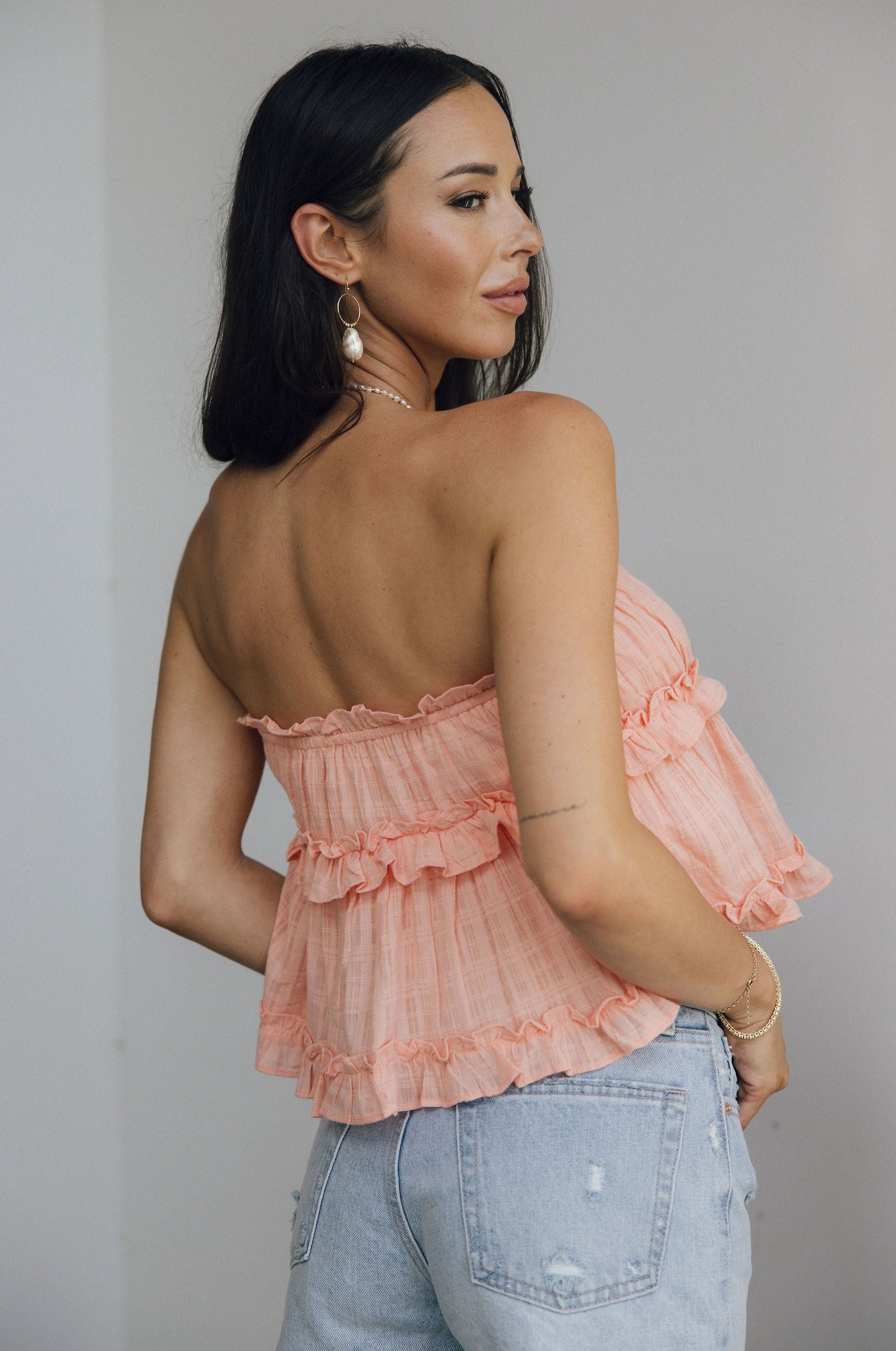 Back view of female model wearing the Millie Peach Ruffle Strapless Top which features Peach Cotton Fabric, Ruffle Tiered Body Details, Peach Lining, Cropped Waist and Strapless