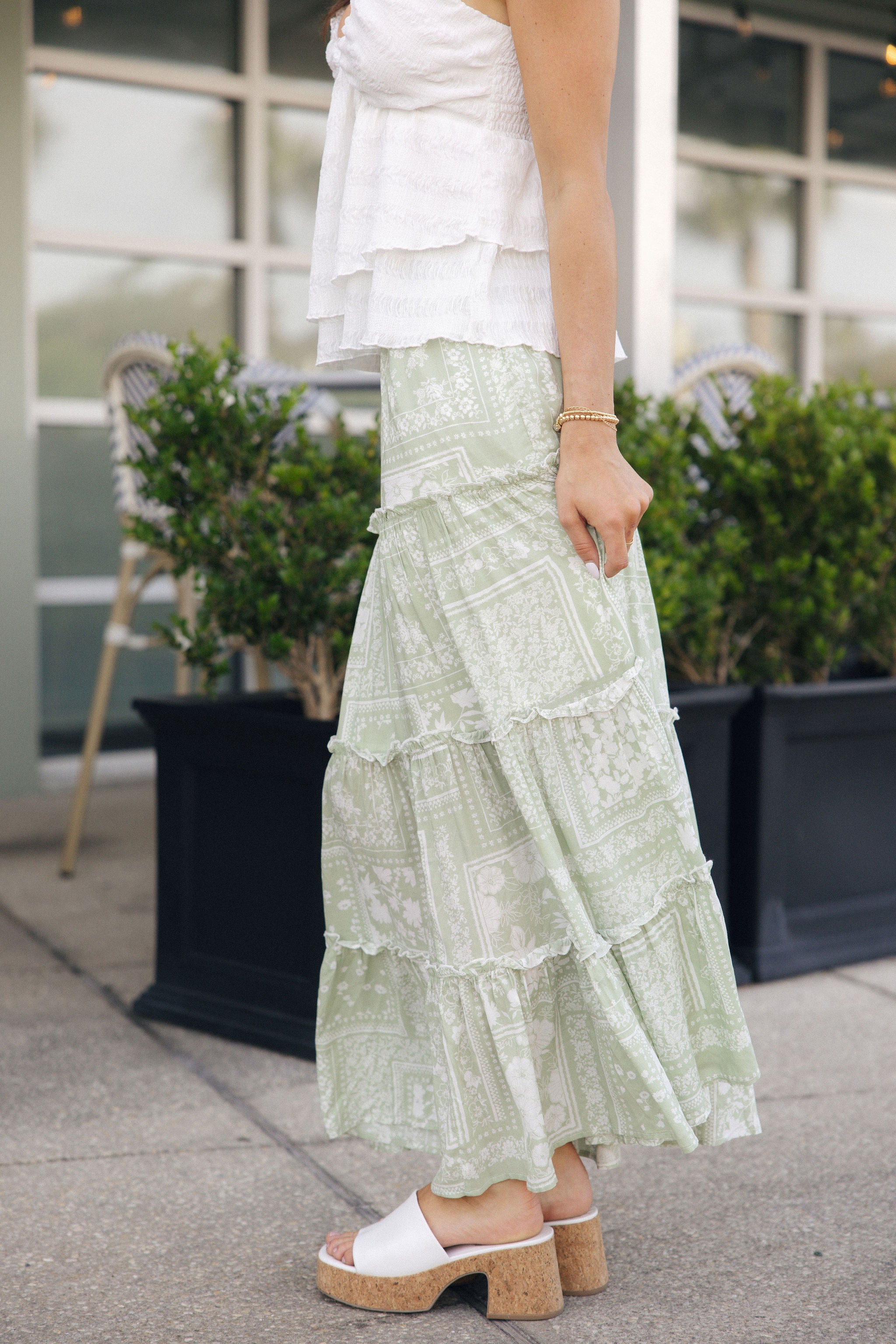 Full body side view of female model wearing the Yasmine Sage & White Floral Maxi Skirt that has sage fabric with a whtite floral patchwork pattern and tiered ruffle details. Paired with white tank top.